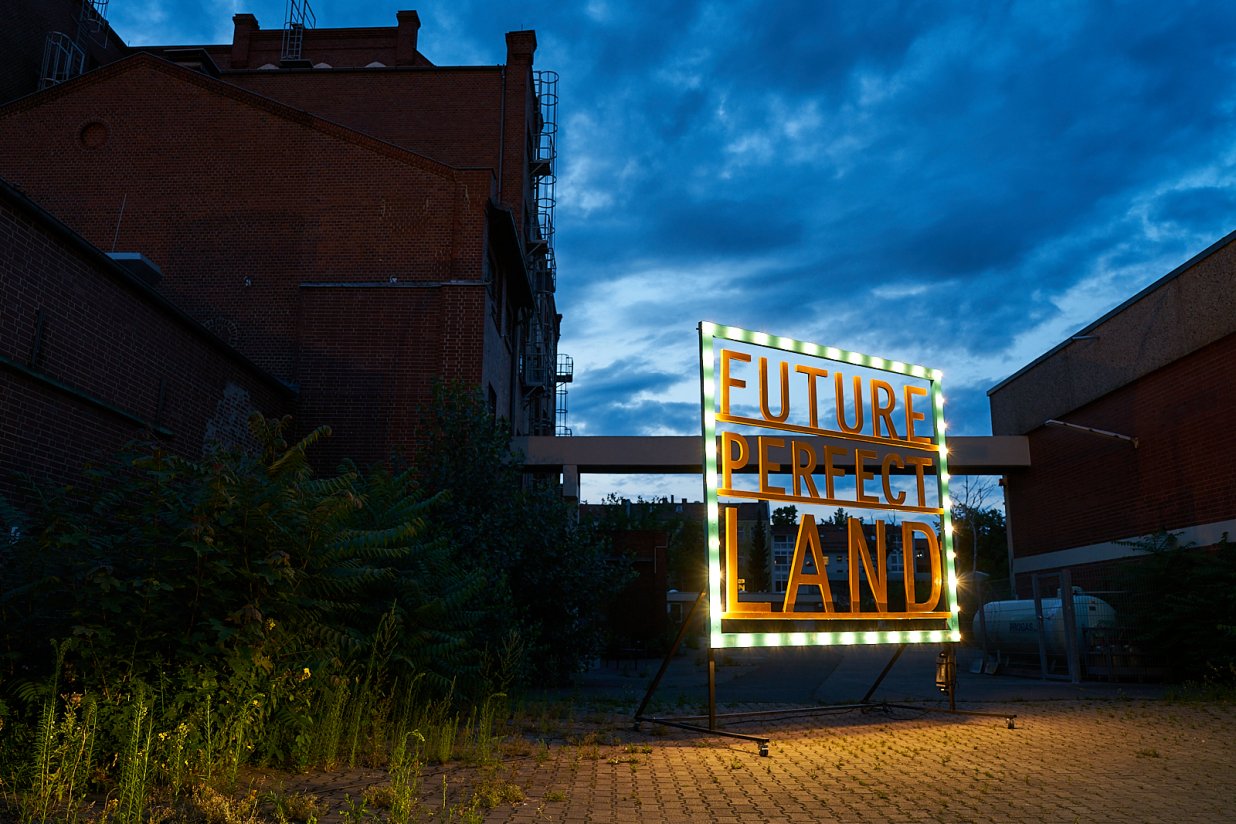 Ein Schild mit der Schrift "FuturePerfectLand" steht auf einem Industriegel&auml;nde, das teilweise von Pflanzen &uuml;berwachsen ist, unter einem drohenden Himmel. | © Pablo Hassmann, Magma Studio