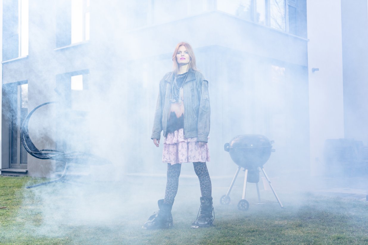 Eine punkig gekleidete Frau steht im Nebel vor einem Einfamilienhaus mit Grill. | © Peter van Heesen