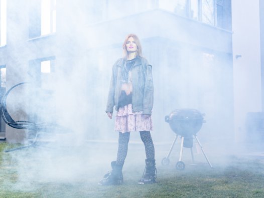 Eine punkig gekleidete Frau steht im Nebel vor einem Einfamilienhaus mit Grill. | © Peter van Heesen
