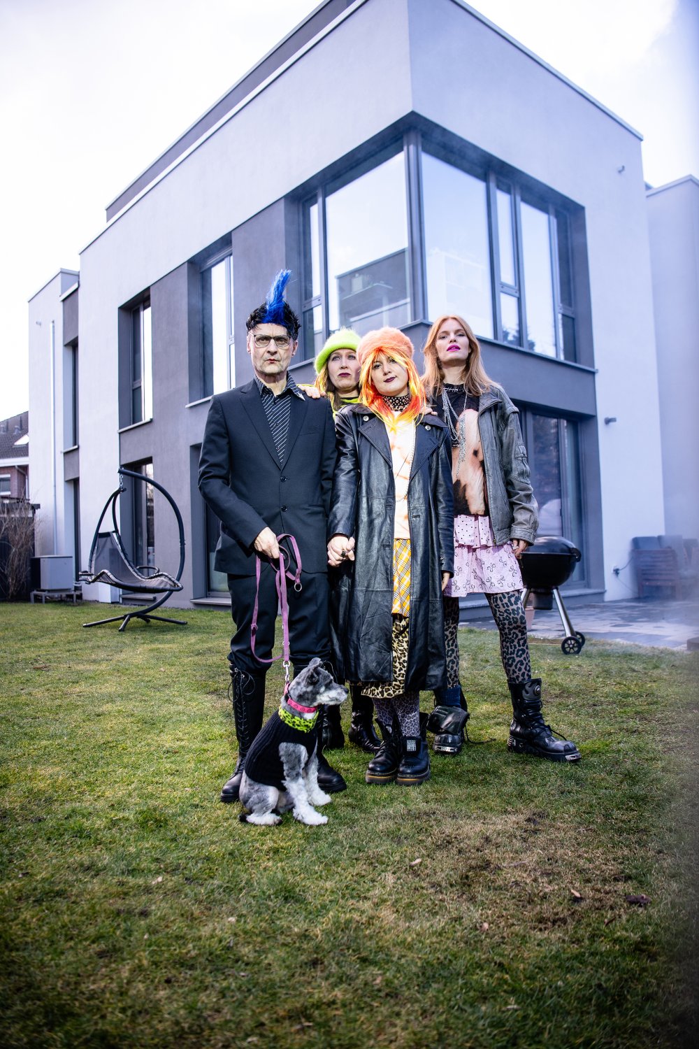Eine punkig gekleidete Gruppe von vier Personen mit Hund steht vor einem schicken Einfamilienhaus. | © Peter van Heesen