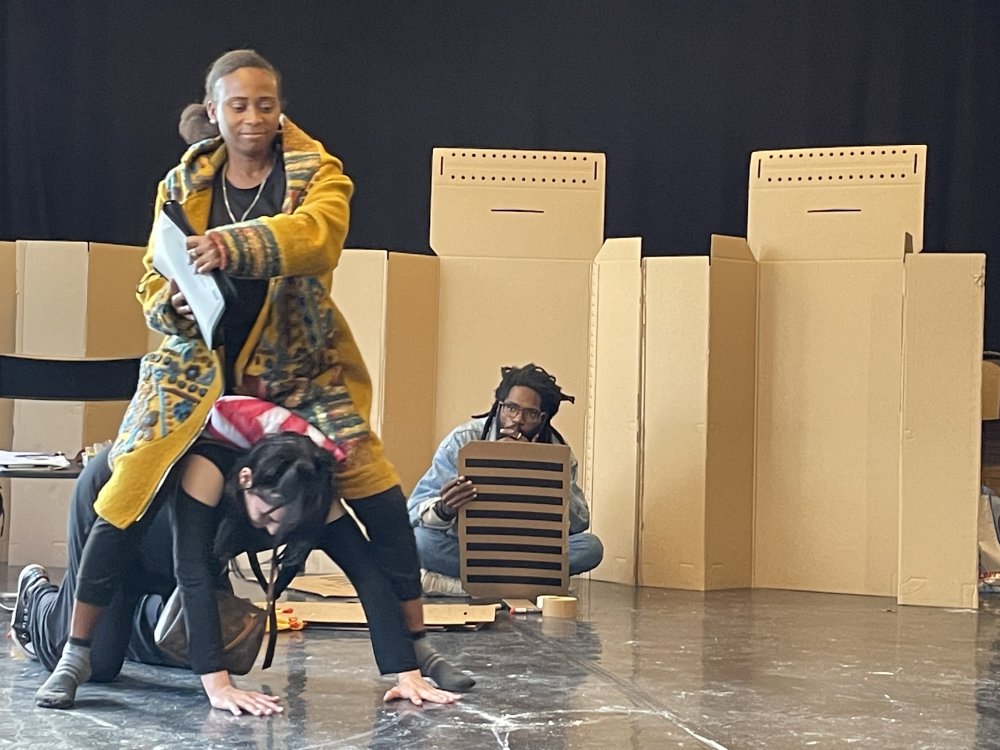 Eine Schauspielerin "reitet" auf einer anderen. Im Hintergund schaut ein weiterer K&uuml;nstler zu. Hinter ihm befindet sich eine Wand aus Pappkartons. | © Carlos Manuel